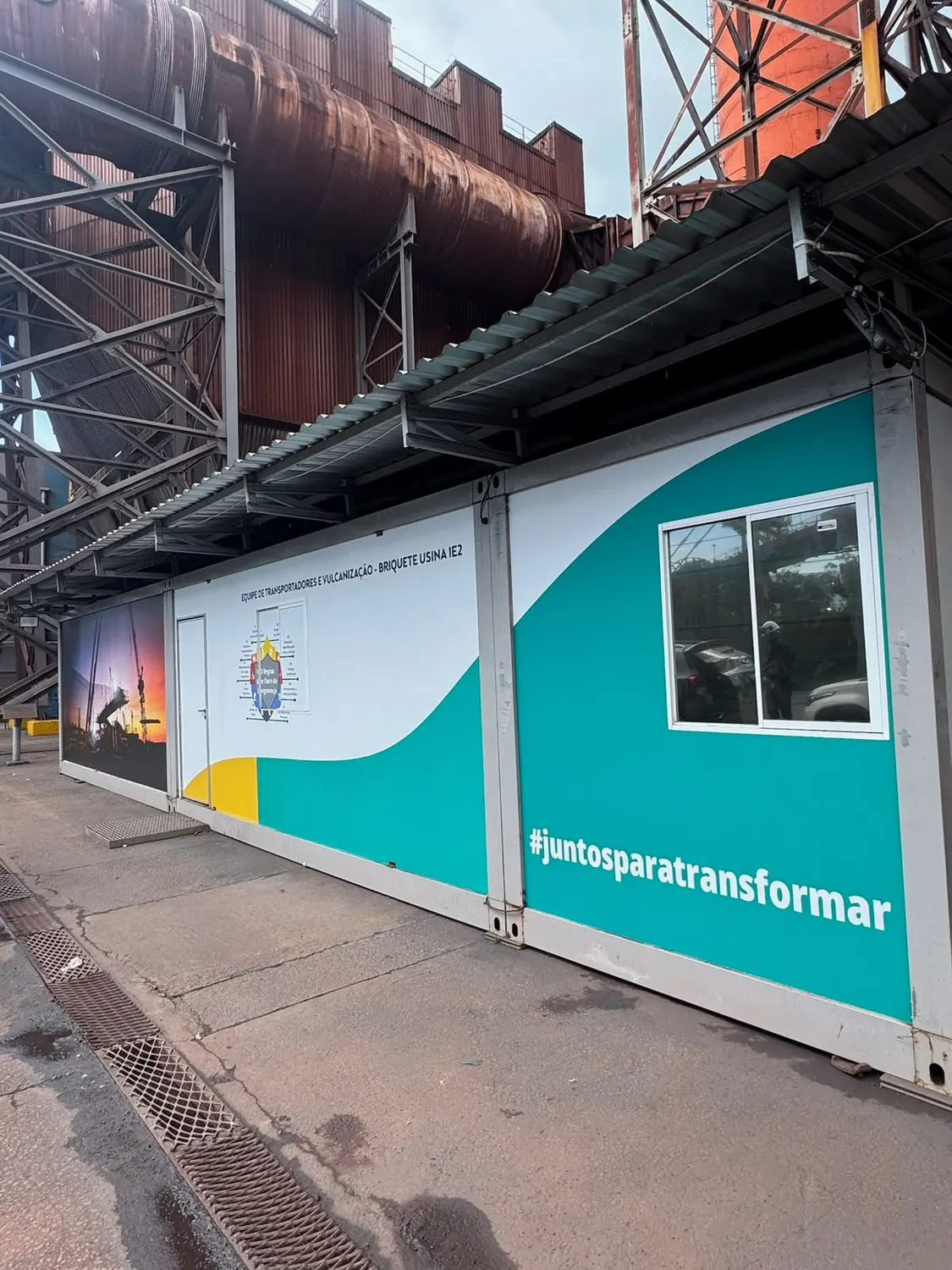 Fileira de containers industriais adesivados em verde-água com a hashtag juntos para transformar
